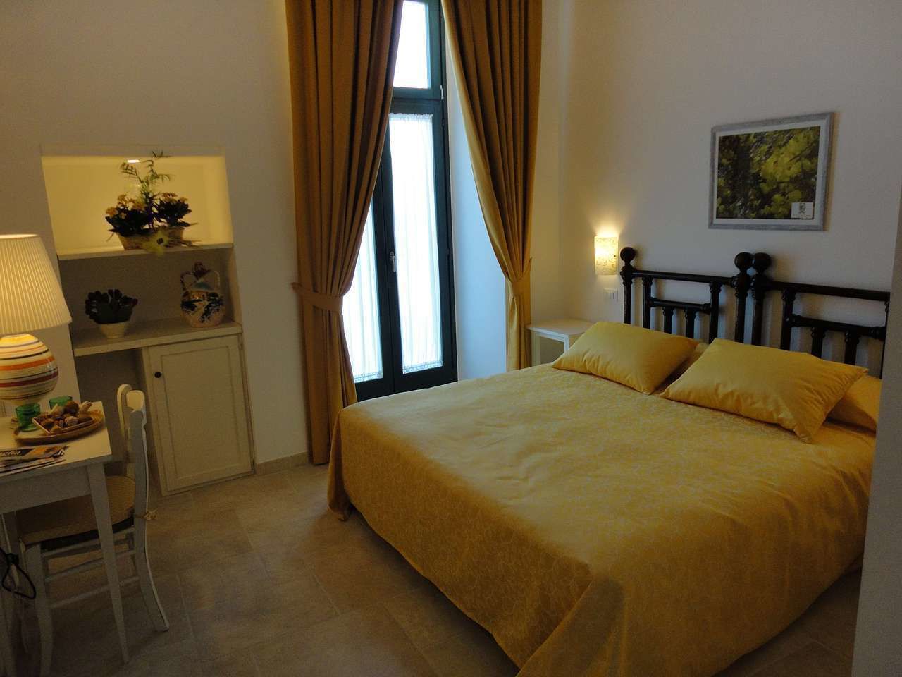 A bedroom with a bed, a desk, and a chair. There is a window with curtains, a lamp, and a picture on the wall.