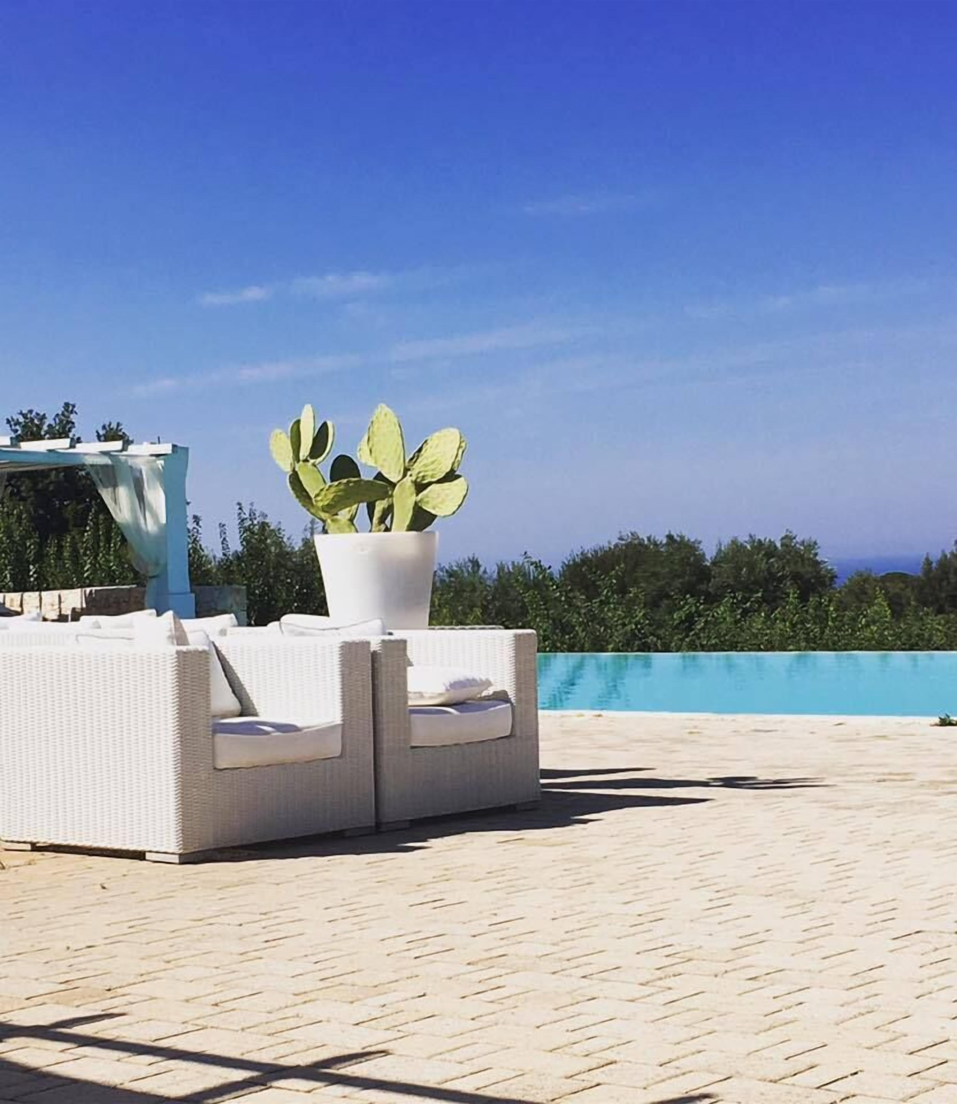 Un área de estar exterior blanca con una planta en maceta, una pérgola y una piscina al fondo.