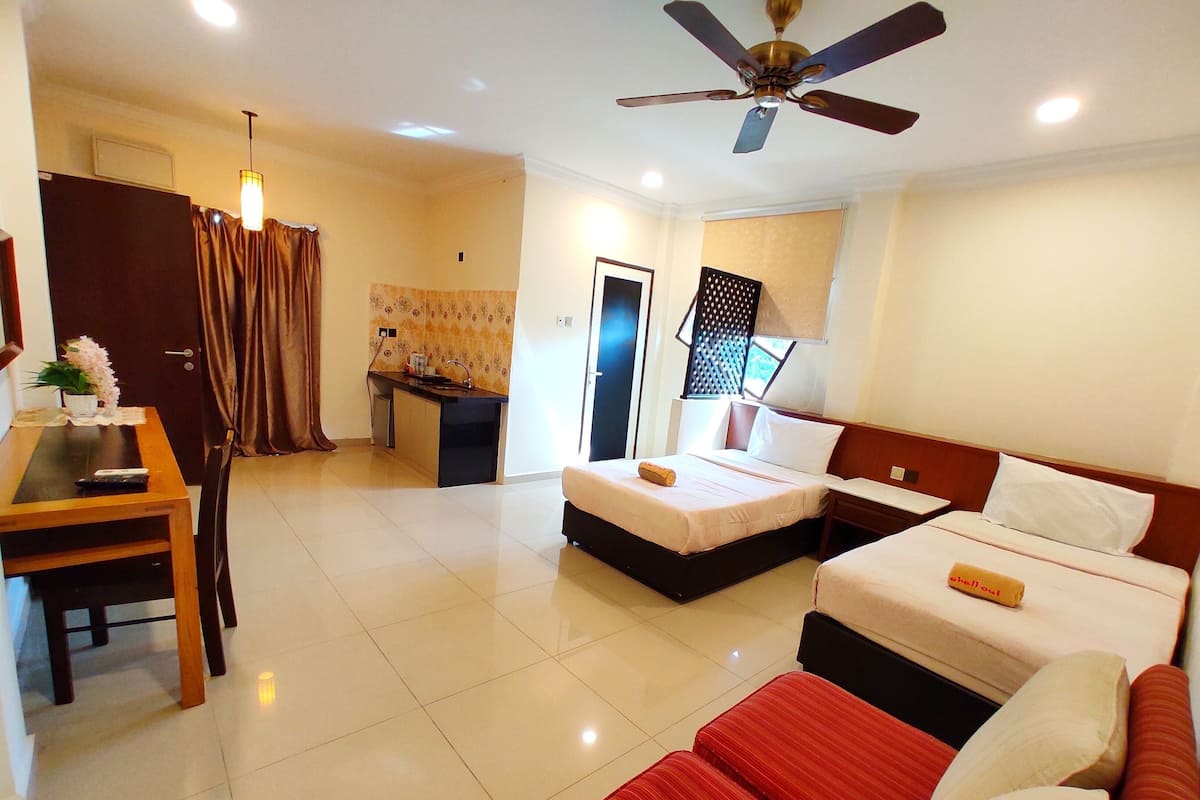A hotel room with two beds, a desk, a chair, a ceiling fan, and a small kitchenette.
