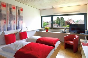 A modern hotel room with a bed, red and white pillows, a red chair, a TV, and a window with a view of houses. - Rotes Haus (Ueberlingen)