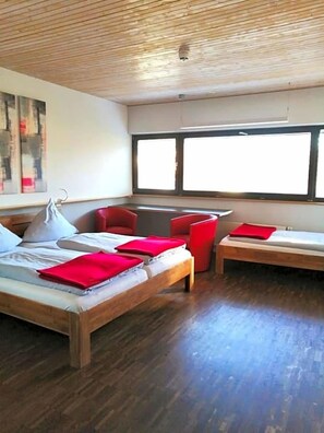 Ett modernt hotellrum med en stor säng, två mindre sängar, en sittgrupp och ett stort fönster som släpper in naturligt ljus.