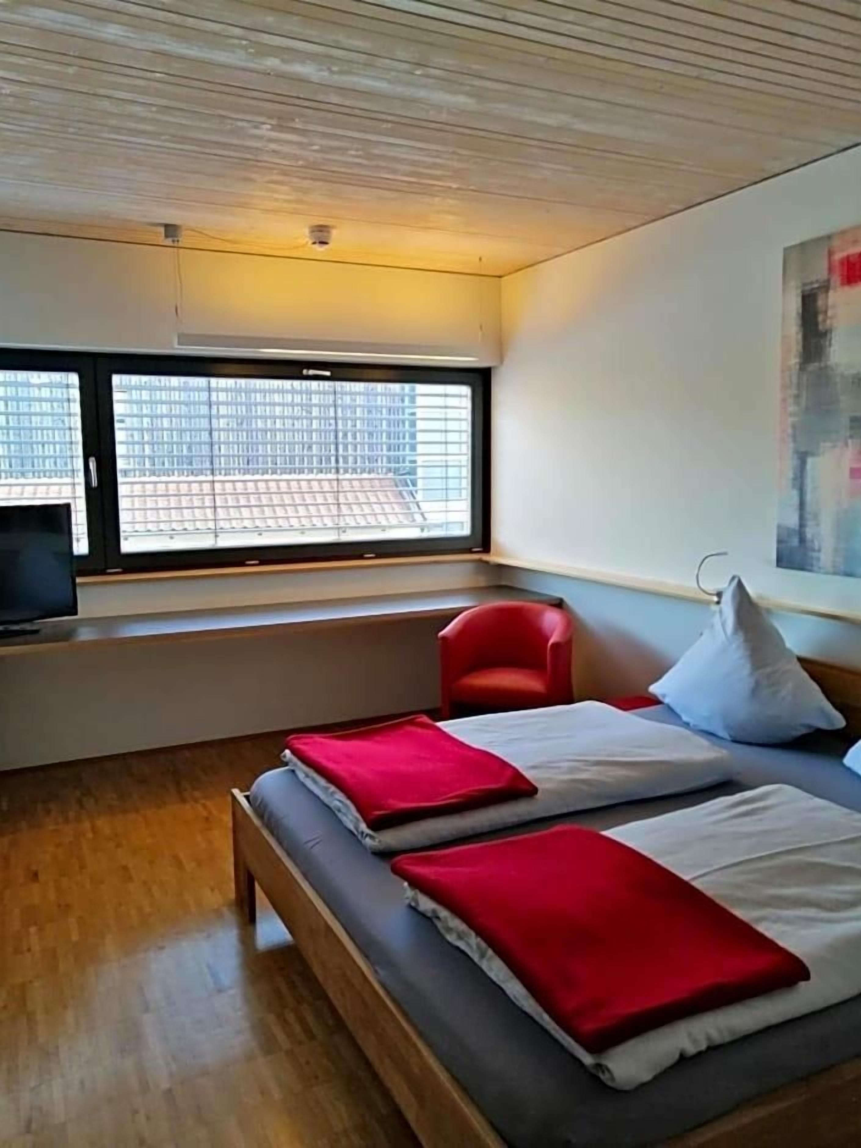 Ein Hotelzimmer mit Bett, einem roten Sessel, einem Fernseher und einem Fenster mit Stadtblick.