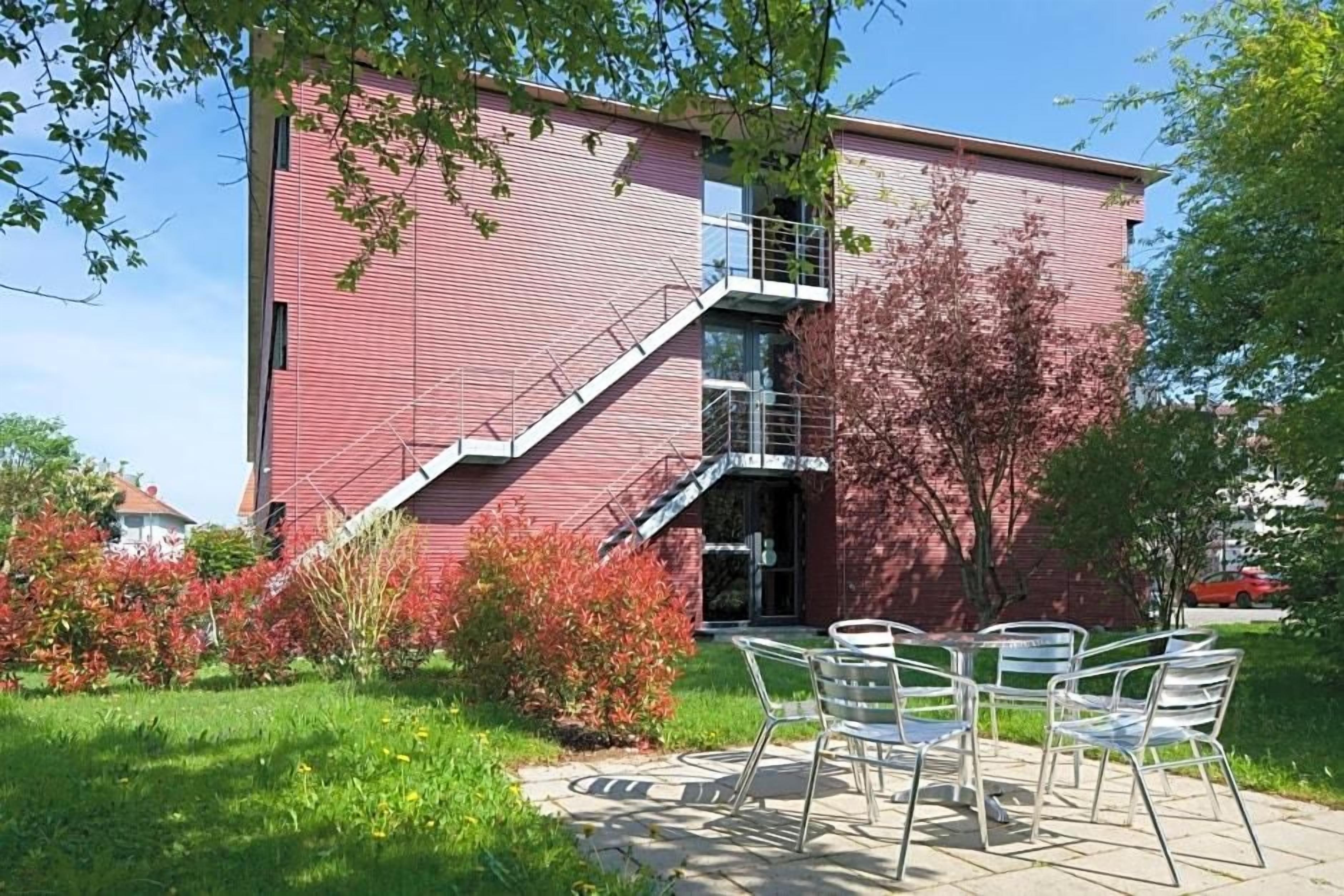 A red brick building with a staircase leading to a balcony, surrounded by greenery and outdoor seating.