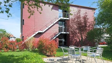 Ein rot backsteiner Gebäude mit einer Treppe, die zu einem Balkon führt, umgeben von Grünpflanzen und Außenmöbeln.
