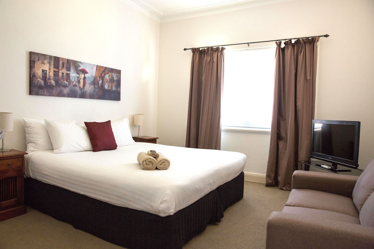 A hotel room with a large bed, a television, a sofa, and a painting on the wall.