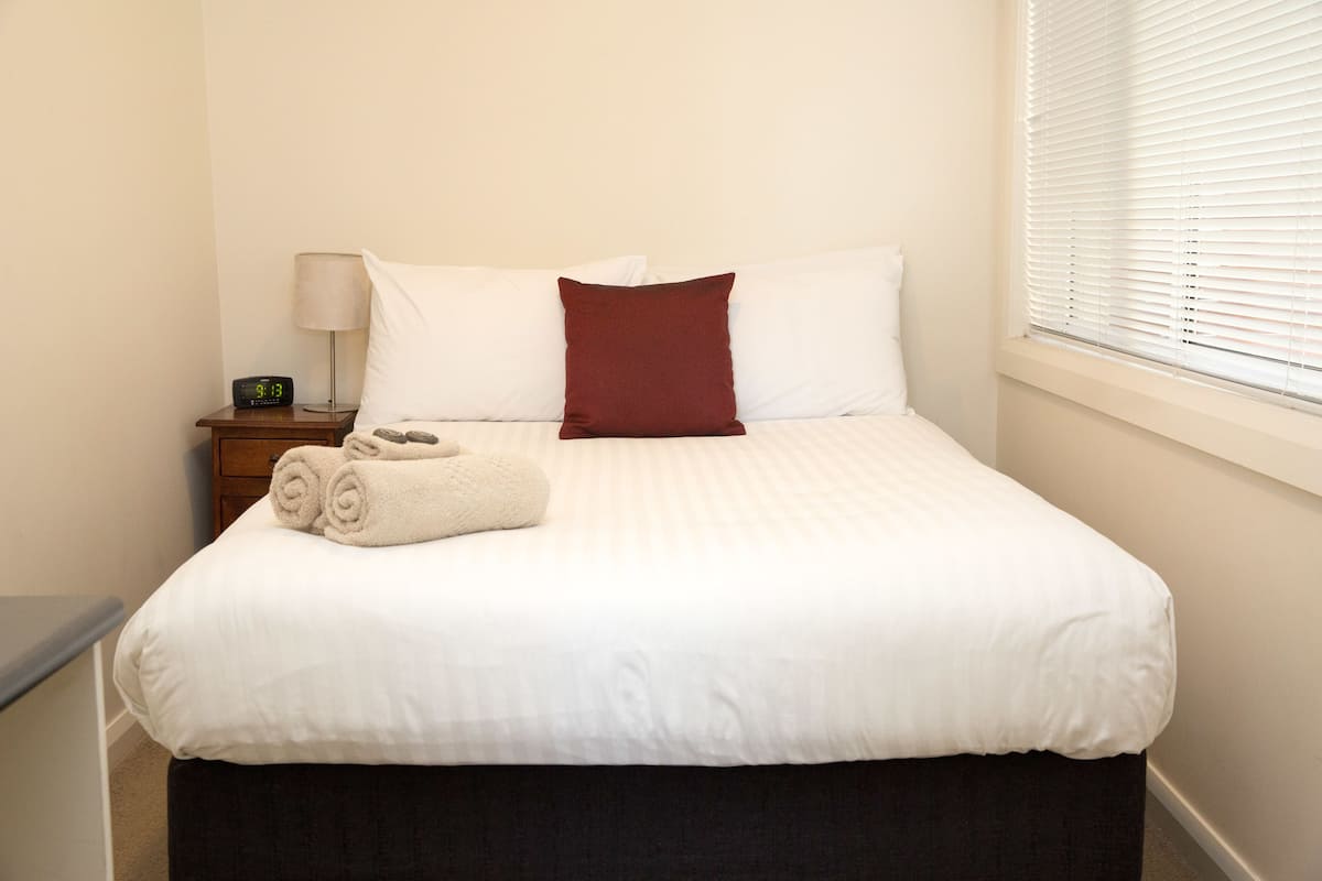 A neatly made bed with white bedding and a single red pillow, a bedside lamp, and a digital clock on a wooden nightstand.