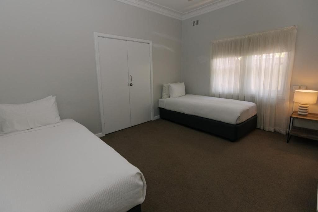 A hotel room with two beds, a wardrobe, a bedside table with a lamp, and a window with curtains.