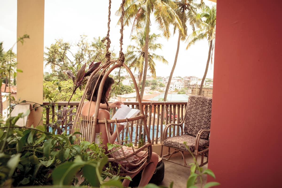 A balcony with a wicker chair and a cushioned chair, overlooking a pool and palm trees.