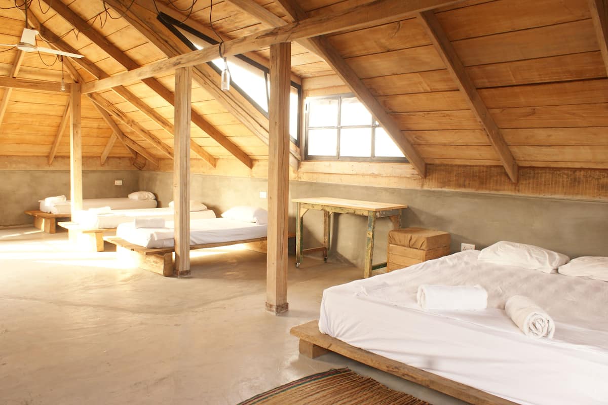 A rustic wooden room with a sloped ceiling, multiple beds, and a large window providing natural light.