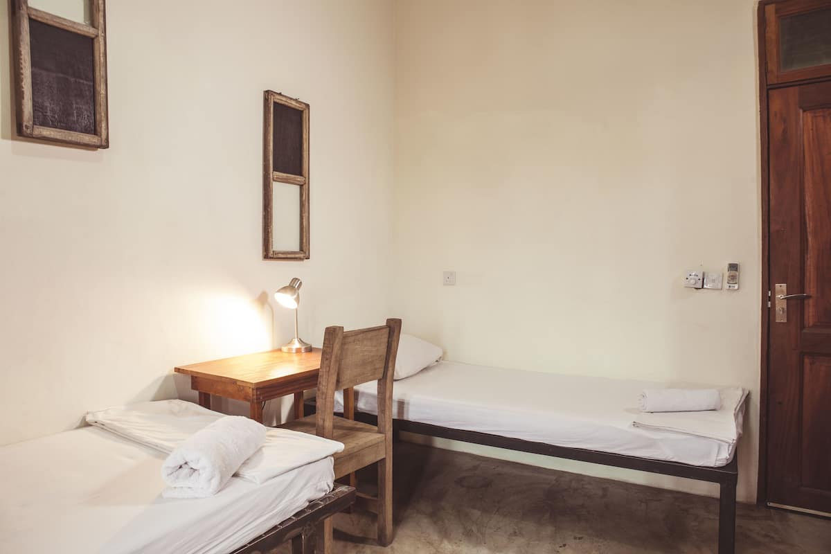A small, simple room with two single beds, a wooden desk with a chair, a desk lamp, and a mirror on the wall.