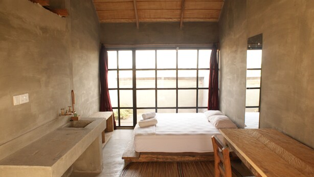 Una habitación con cama, una mesa de madera, un banco de hormigón, una ventana con cortinas y un lavabo.