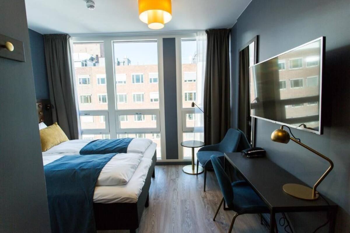 A hotel room with a bed, a desk, a chair, a lamp, and a large window with a view of buildings.