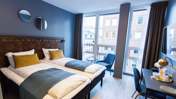 A hotel room with two beds, a dining table, chairs, a small table with a lamp, and large windows offering a view of the city. - Hotel Verdandi Oslo (Oslo)