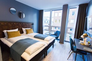 Habitación de hotel con dos camas, una mesa de comedor, sillas, una mesita con lámpara y ventanas amplias con vistas a la ciudad.
