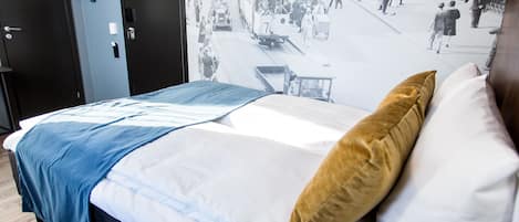 Una cama con colcha azul y blanca, un almohadón marrón y una pared con una escena callejera antigua.