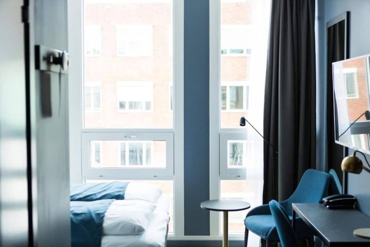 A hotel room with a bed, a desk, a chair, a lamp, and a window with a view of buildings.