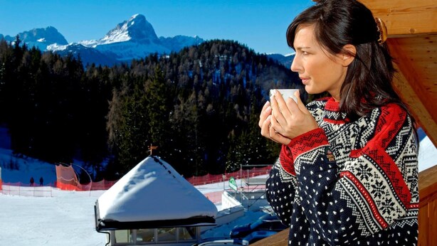 A person in a patterned sweater holding a cup, with a snowy mountain and forest in the background. - Hotel Jú Furcia (Marebbe)