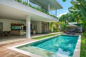 A modern outdoor pool area with a clear pool, wooden deck, and a dining area with chairs and a table. - Villa Paloma (Seminyak)