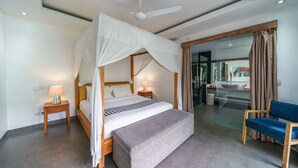 Un dormitorio con cama con dosel, una mesita de noche con lámpara, un banco, una silla y vista al baño a través de un vidrio.