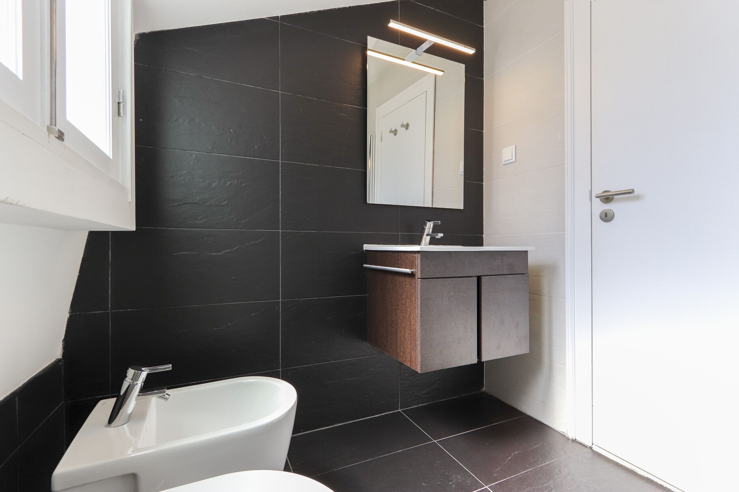 Ein modernes Badezimmer mit einem weißen Waschbecken, dunkel gefliesten Wänden und einem großen Spiegel.