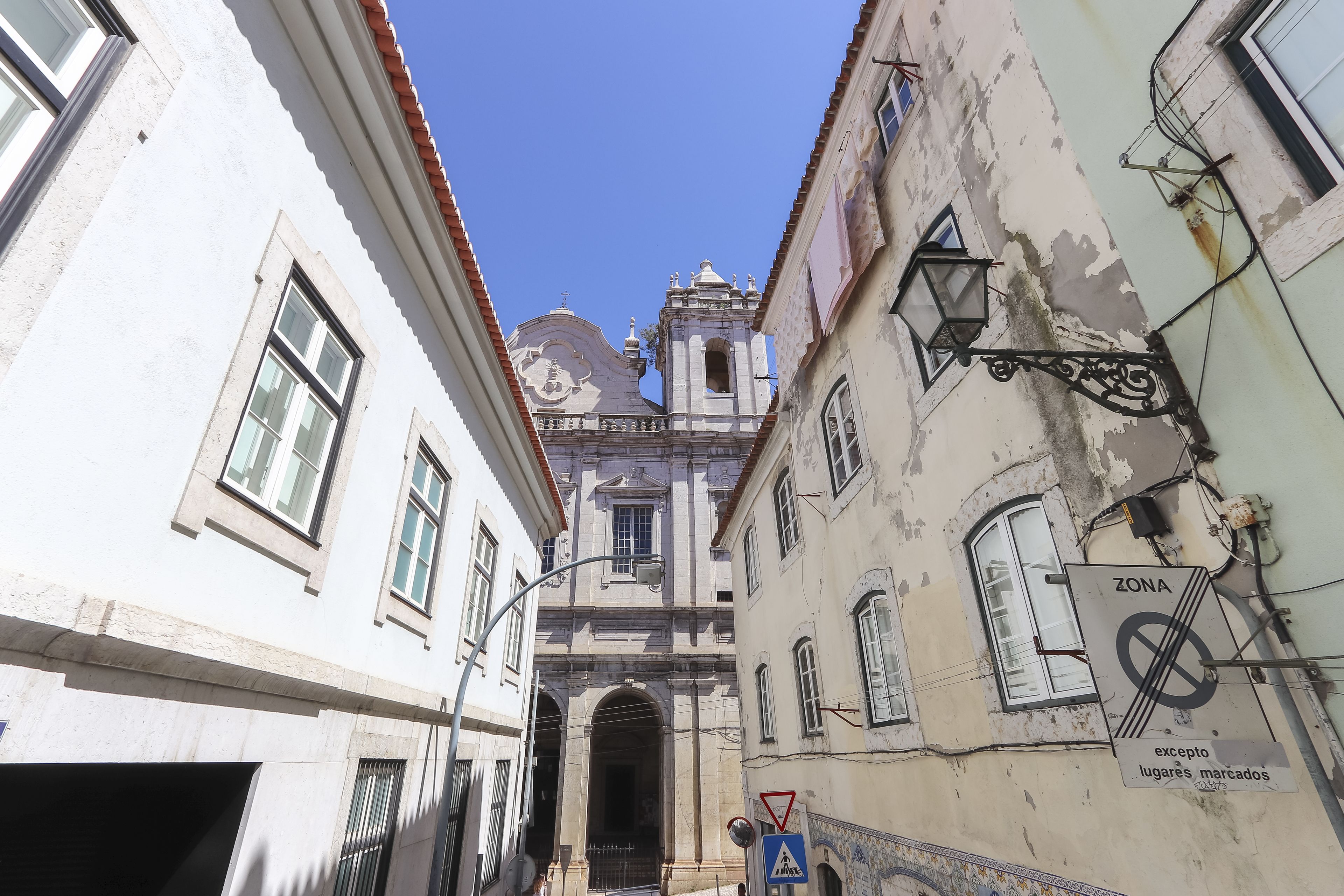 A narrow street with historic buildings, a church with a bell tower, and a 'Zona' sign indicating a restricted area.