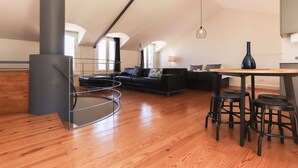 Ein großzügiges Loft mit hohen Decken, Holzboden, einer schwarzen Leder-Couch, einem Esstisch mit Hockern und einem großen Fenster, das für natürliches Licht sorgt.