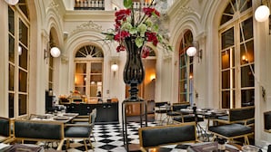 A hotel lobby with a checkered floor, black and gold chairs, a central vase with flowers, and a reception desk in the background. - Hotel Lobby  (Seville)