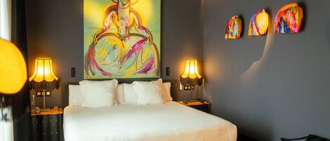 A modern hotel room with a large bed, two bedside tables with lamps, a ceiling fan, and a large abstract painting on the wall.