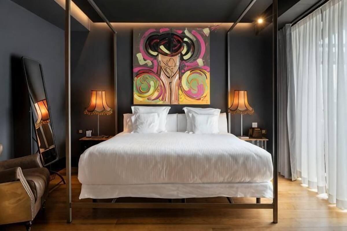 Moderni hotellihuone, jossa on suuri sänky, kaksi yöpöytävalaisinta, seinätaideteos ja suuri ikkuna, jossa on verhot.