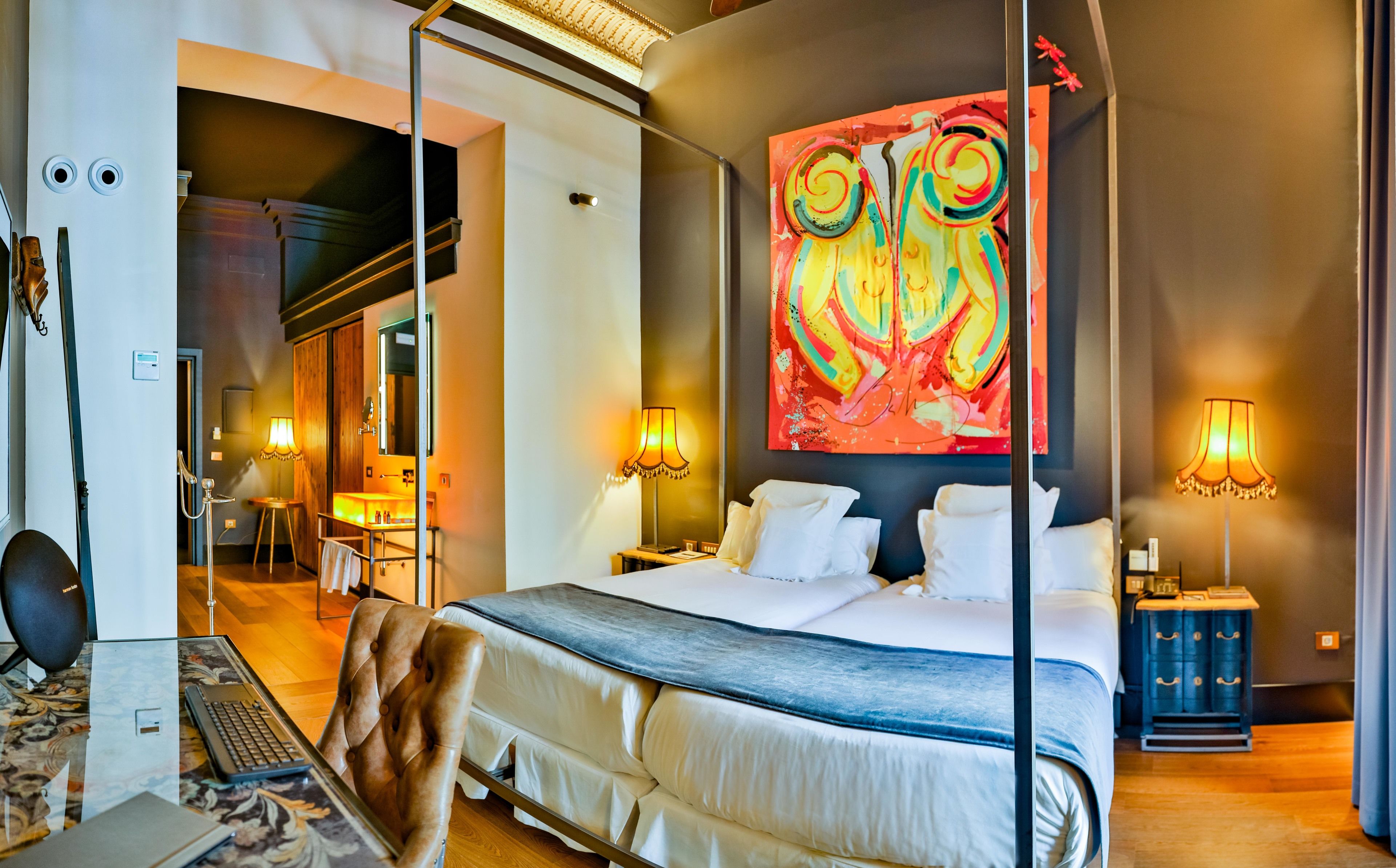 A modern hotel room with a large bed, two bedside lamps, a large abstract painting, and a view of the hallway.