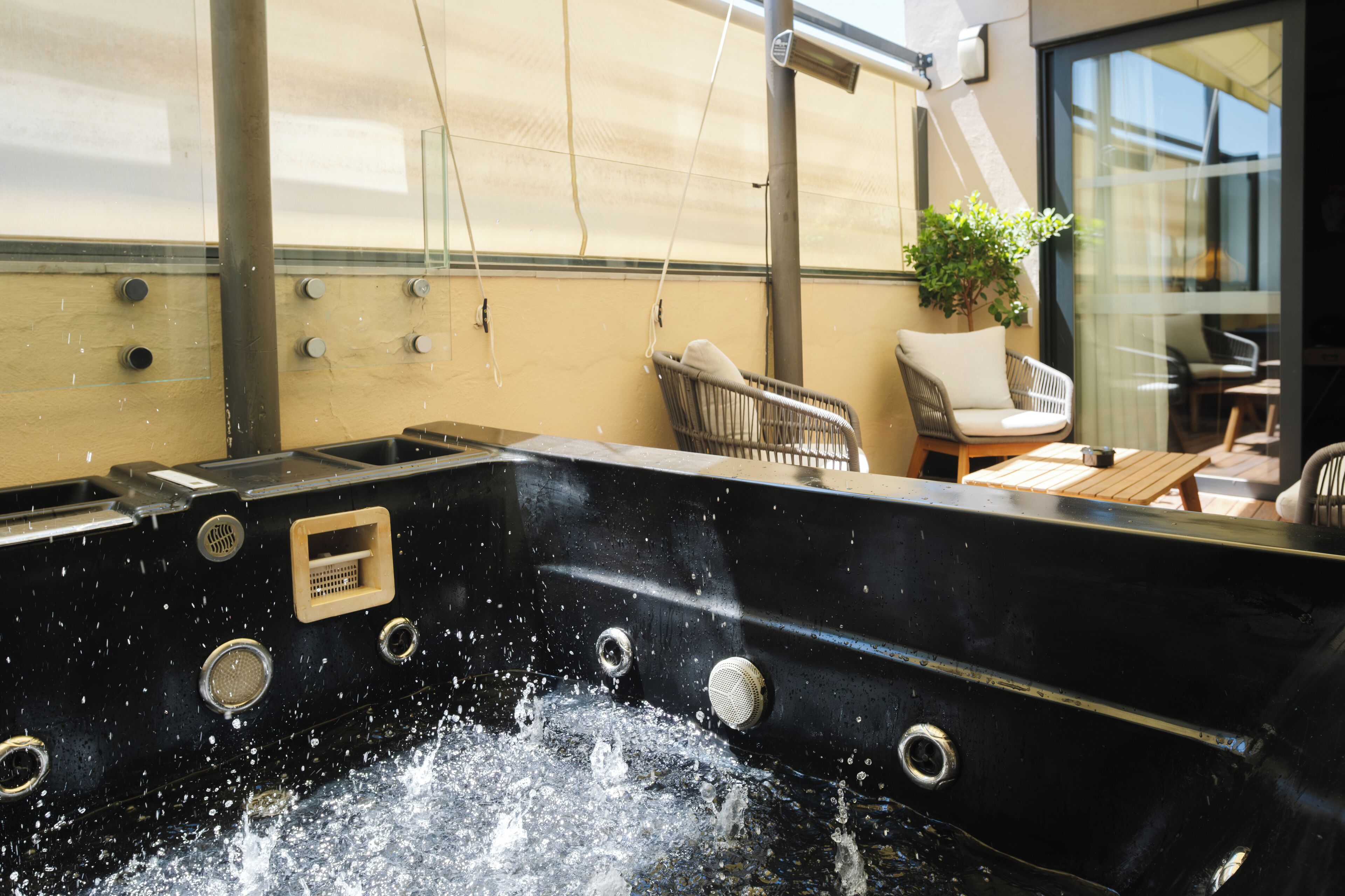 Jacuzzi con chorros de agua, una silla y una mesa en una habitación con pared de vidrio y una planta.