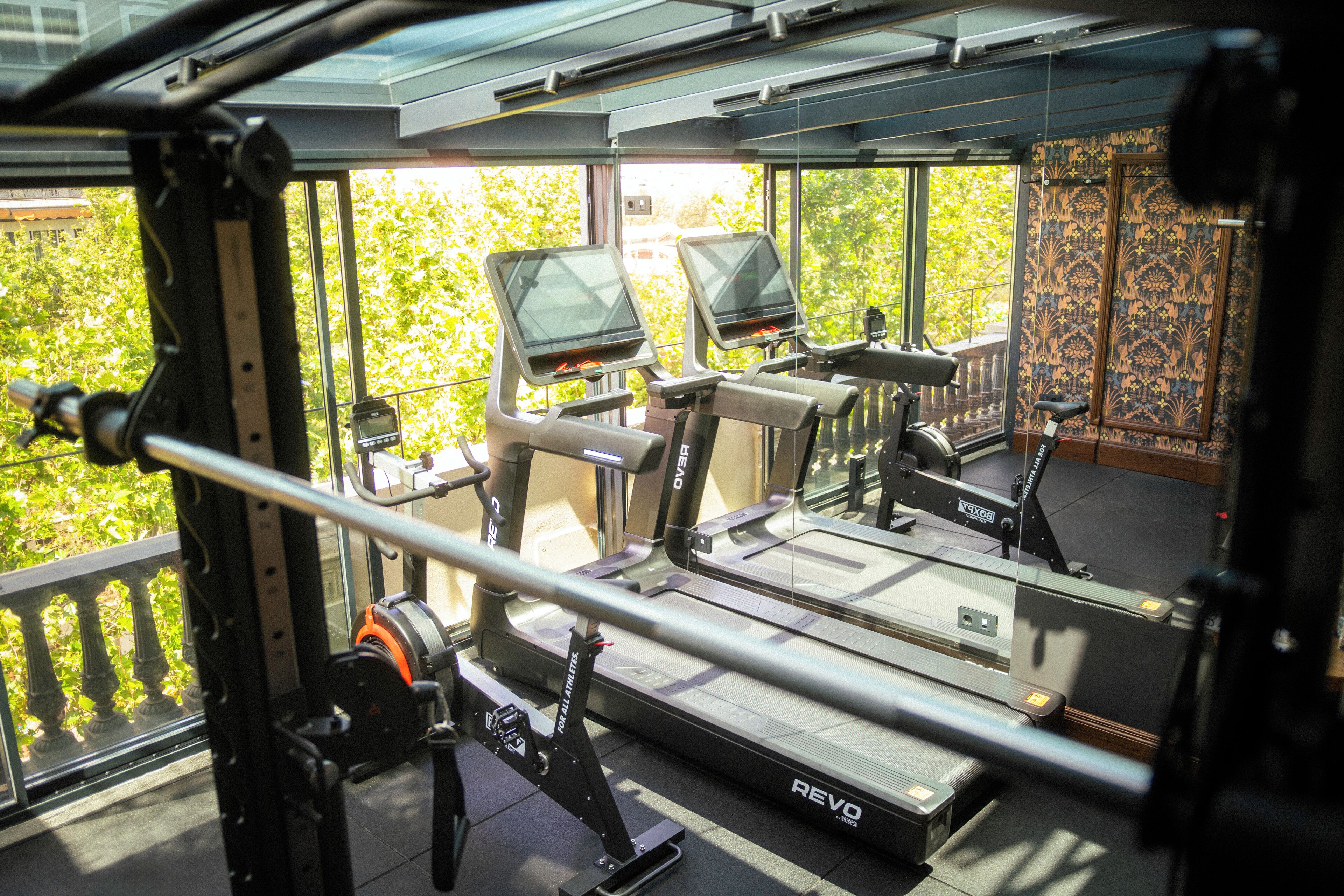 Uma academia com esteiras, máquinas de musculação e uma grande janela com vista para uma área verde.