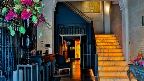 A grand staircase with a warm glow, leading to a dark hallway. A large vase with pink flowers is prominently displayed on the left. - Hotel Lobby  (Seville)