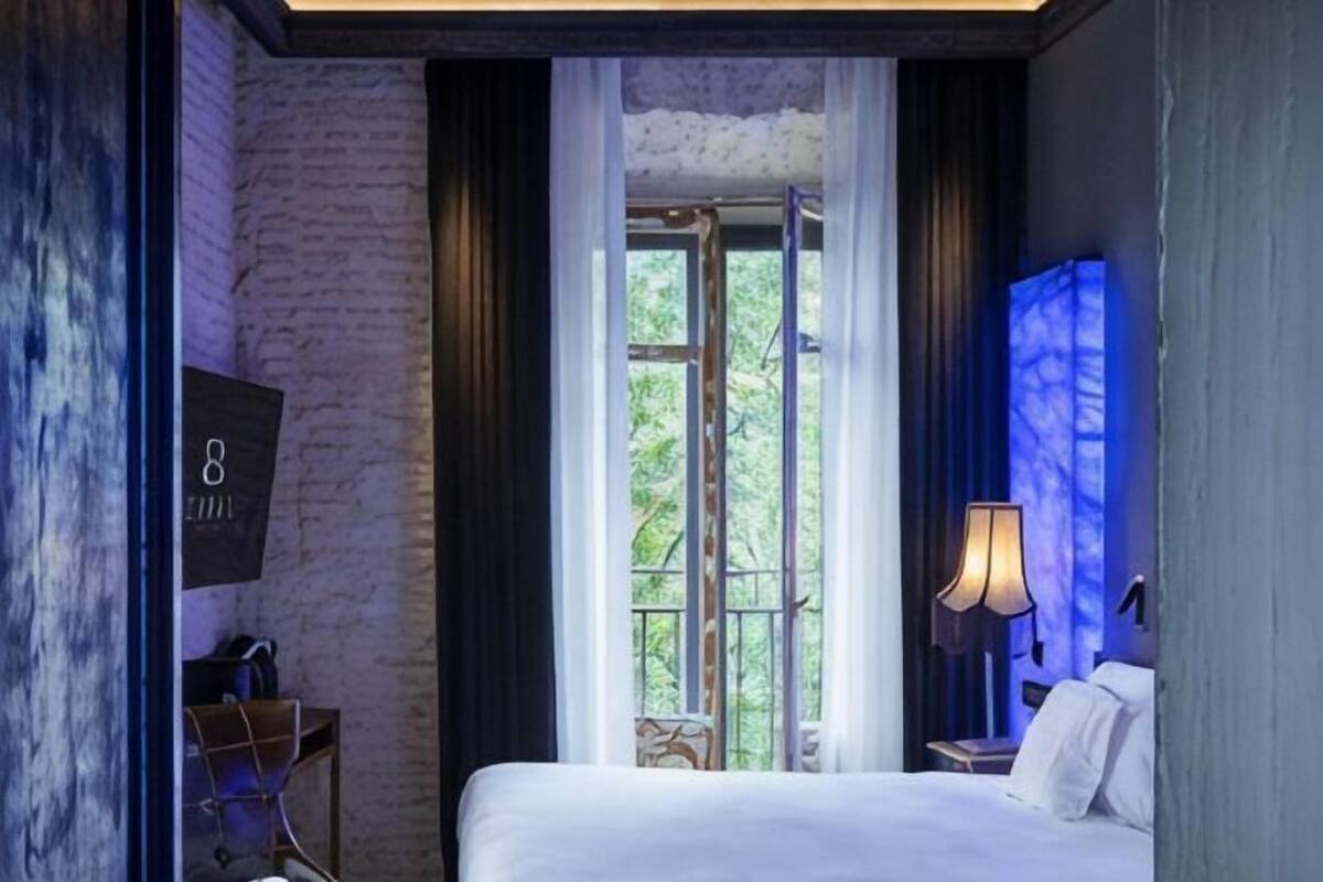Hotellihuone, jossa on suuri sänky, ikkuna, jossa on verhot, lamppu ja seinälle asennettu televisio.