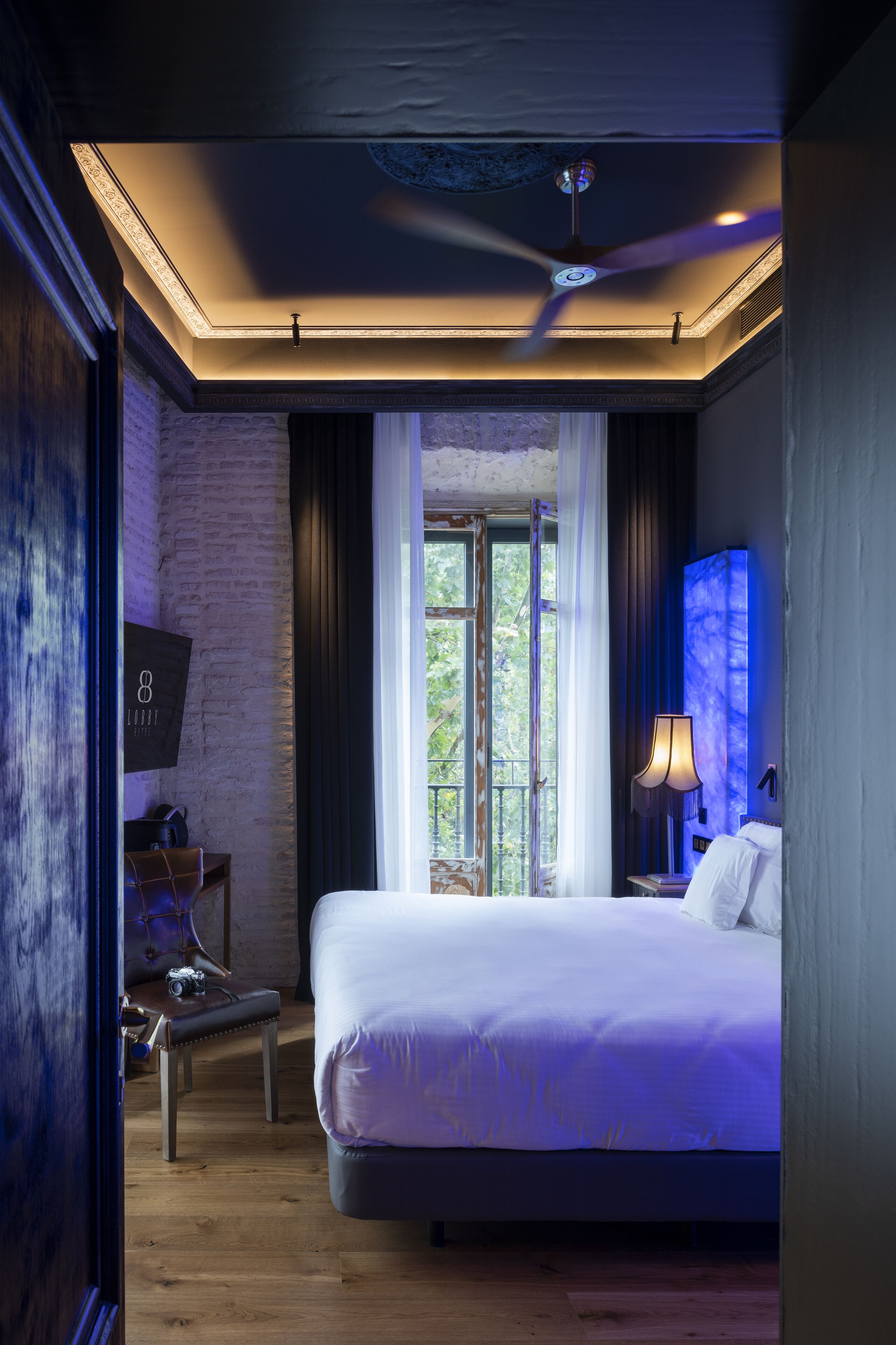 A modern hotel room with a large bed, a ceiling fan, a window with curtains, and a nightstand with a lamp.