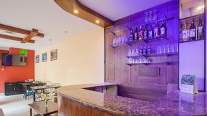 A modern bar area with a granite countertop, glass shelves with bottles and glasses, a flat-screen TV on the wall, and a dining area with a table and chairs.
