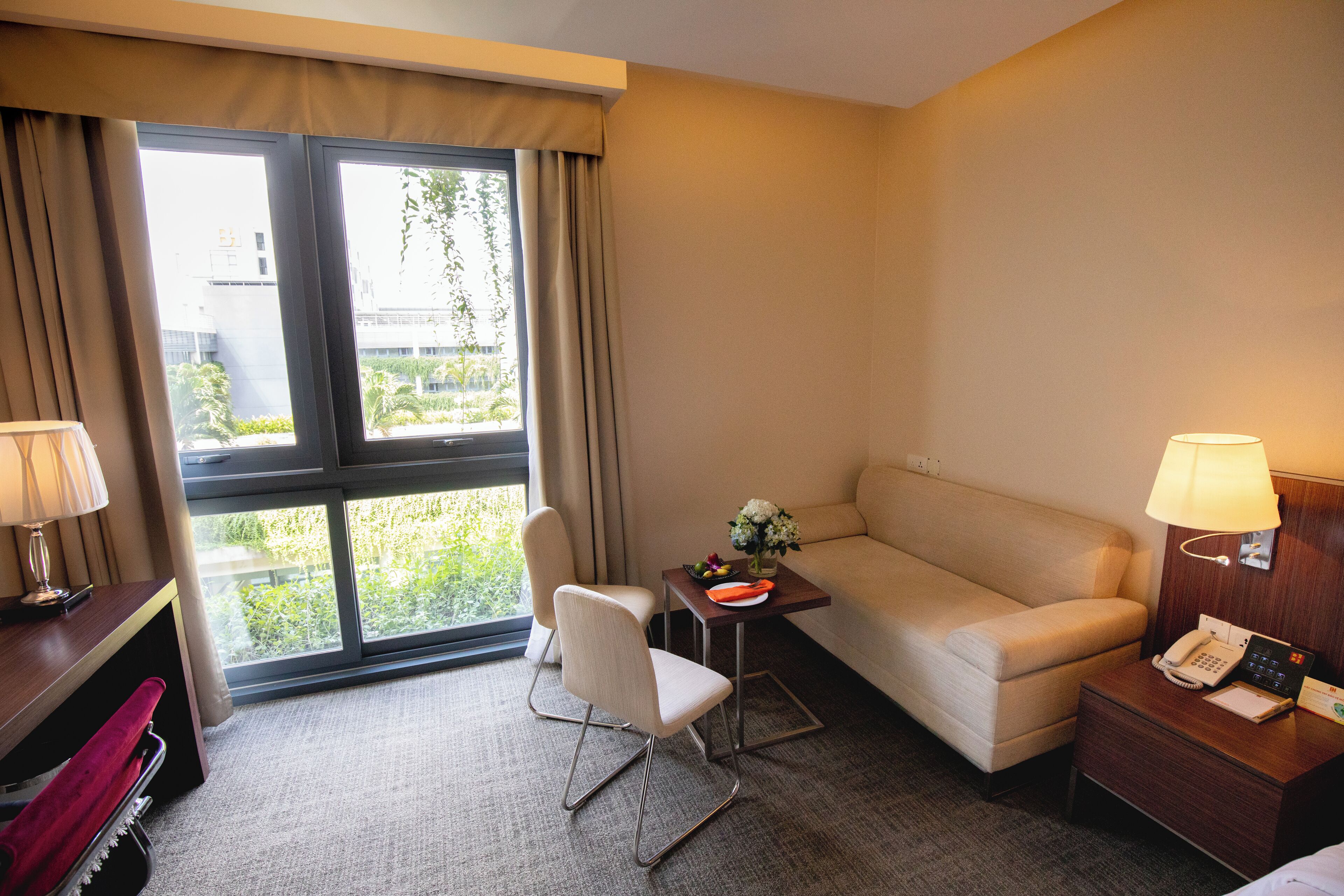 A hotel room with a sofa, a chair, a small table with a lamp, a bedside table with a phone, and a window with curtains.