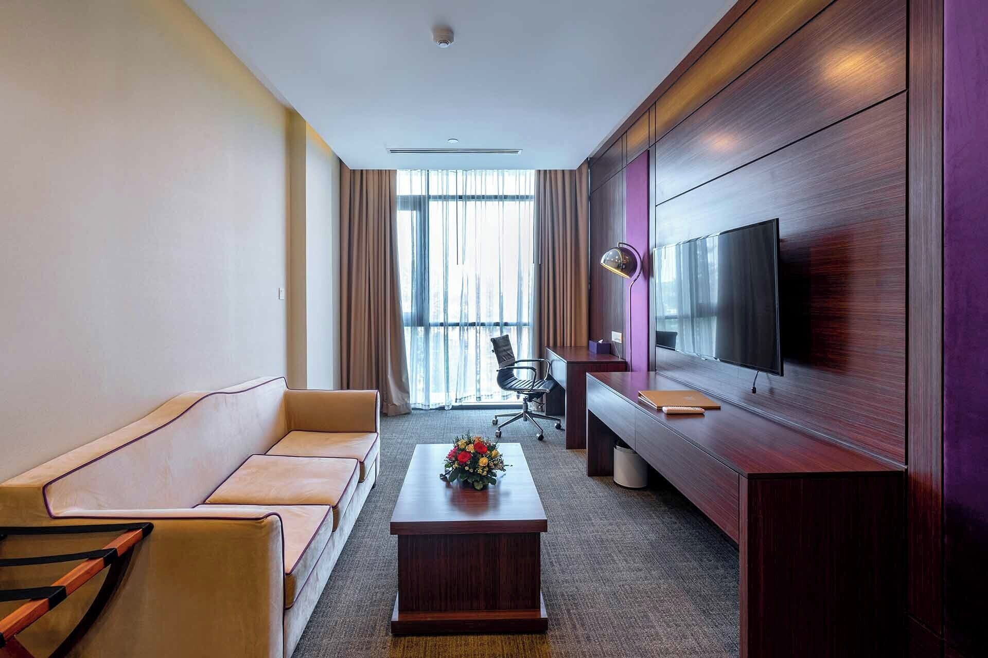 A hotel room with a sofa, a coffee table with flowers, a desk with a computer, and a TV on the wall.