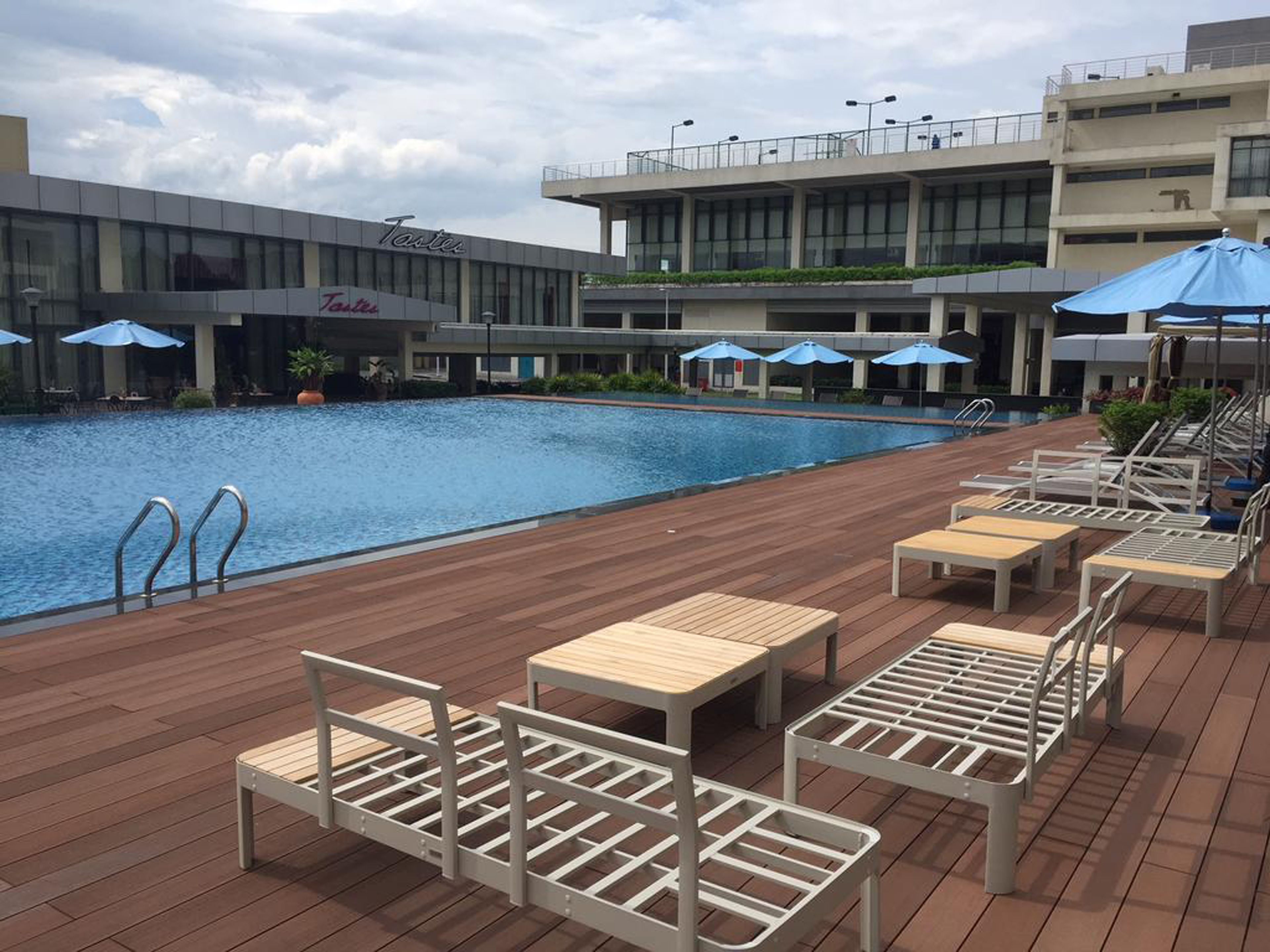 A poolside area with lounge chairs and umbrellas, a building with 'Taste' signage, and a clear sky.
