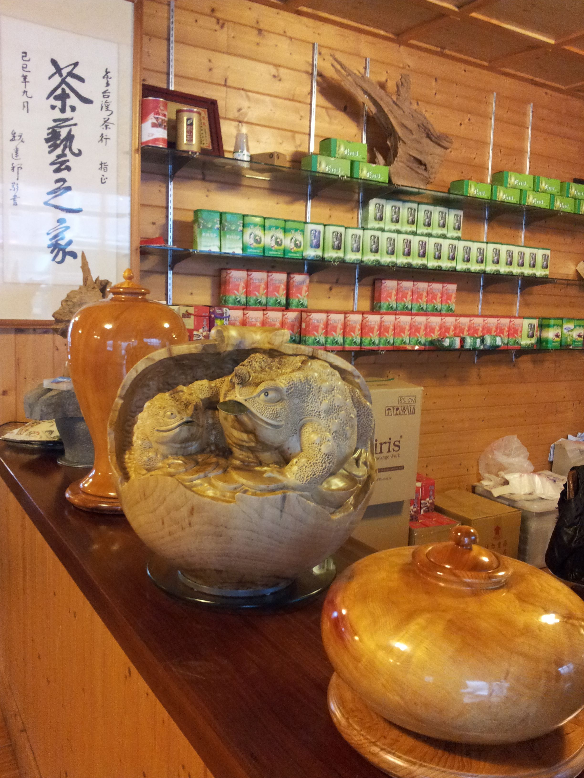 A wooden interior with a counter displaying various items including a large ceramic fish sculpture, a wooden urn, and shelves stocked with green and red packaged goods.