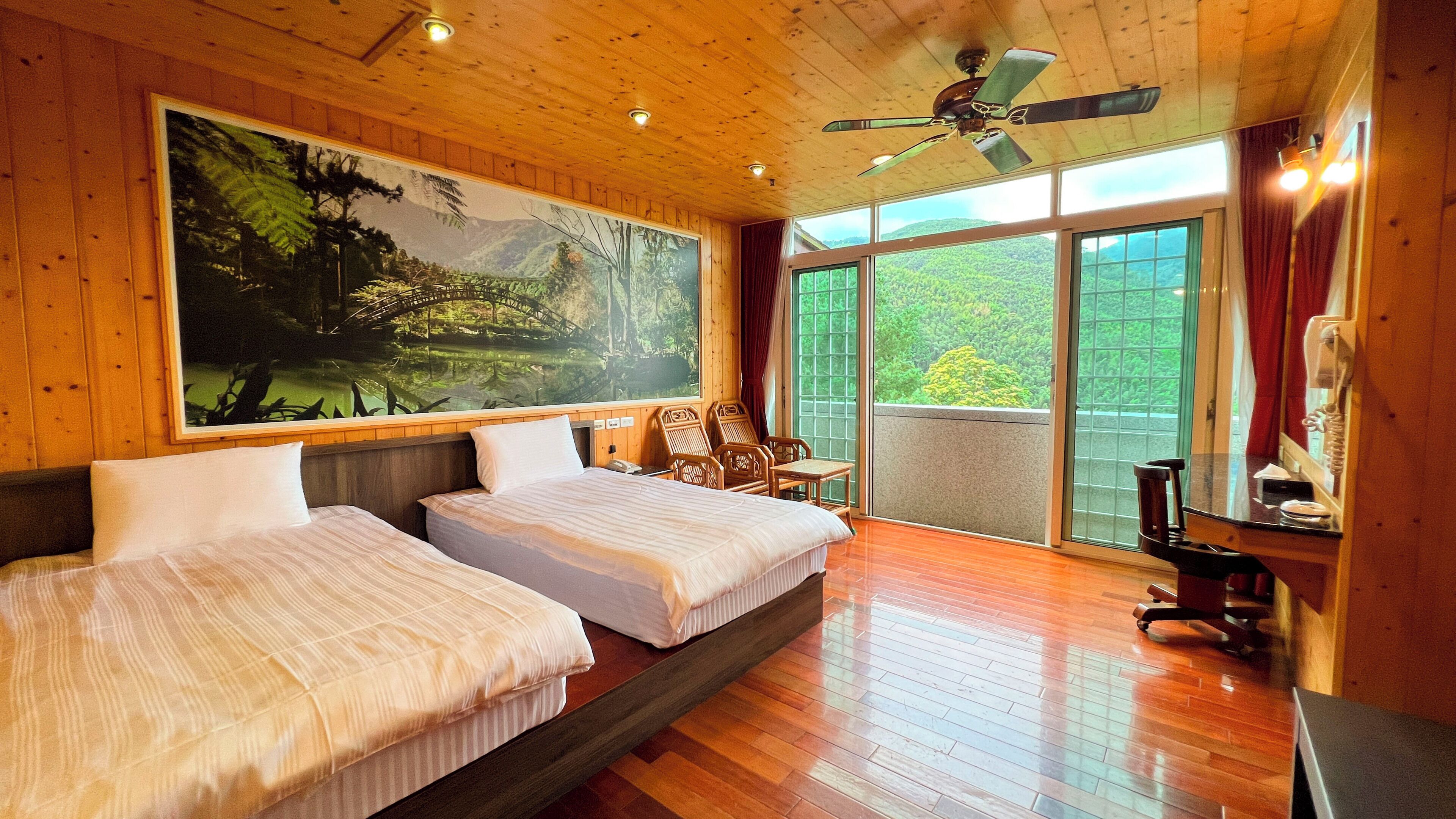 Een kamer met twee bedden, een groot raam met uitzicht op groen en een plafondventilator.