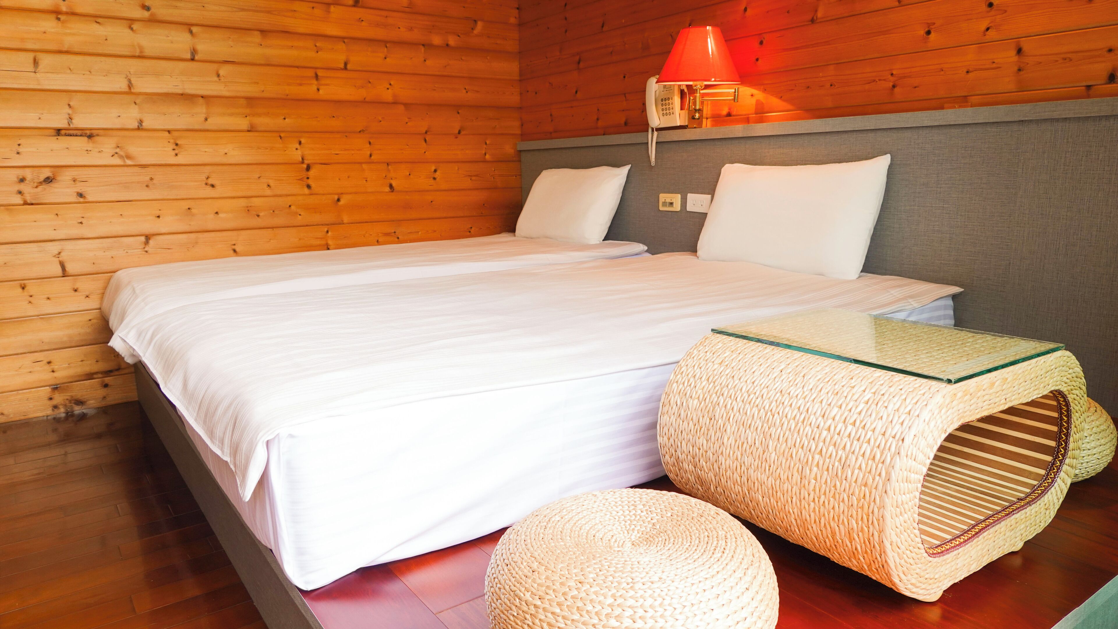 A wooden cabin-style room with a bed, a woven pouf, and a red lamp on the wall.