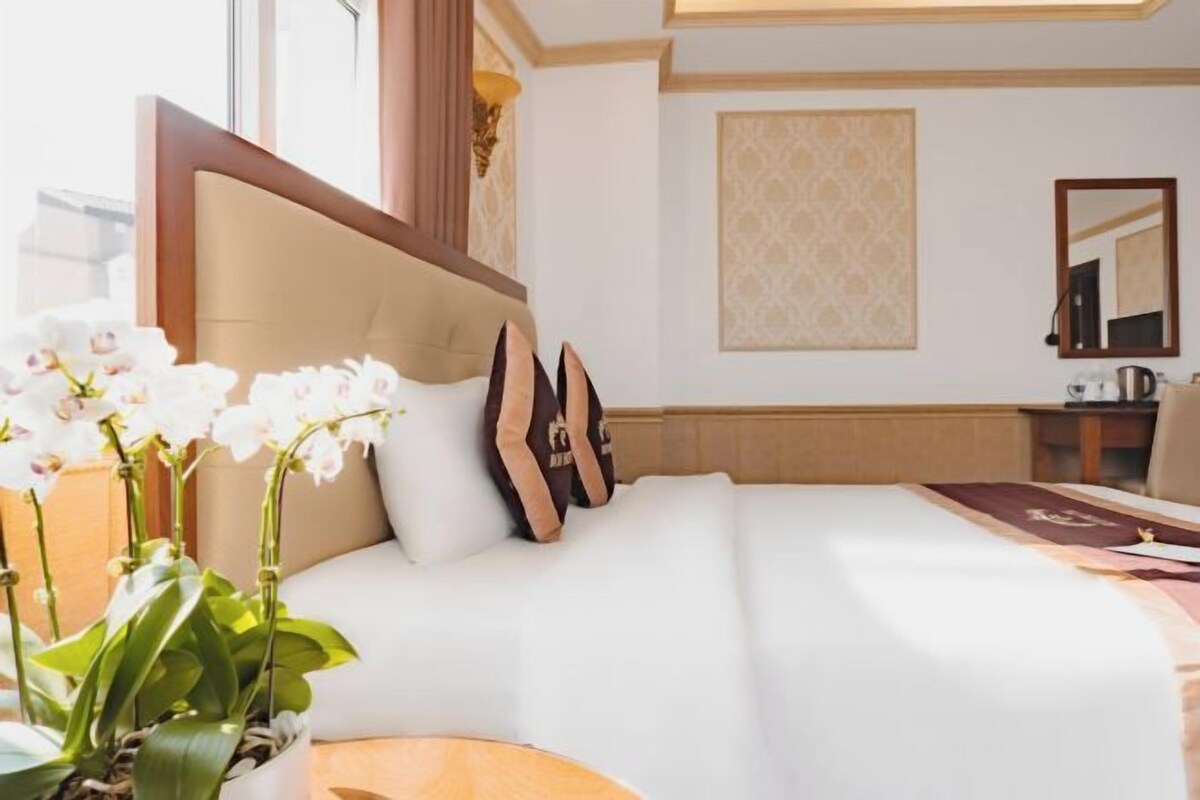 A hotel room with a large bed, a bedside table, a mirror, and a potted plant.