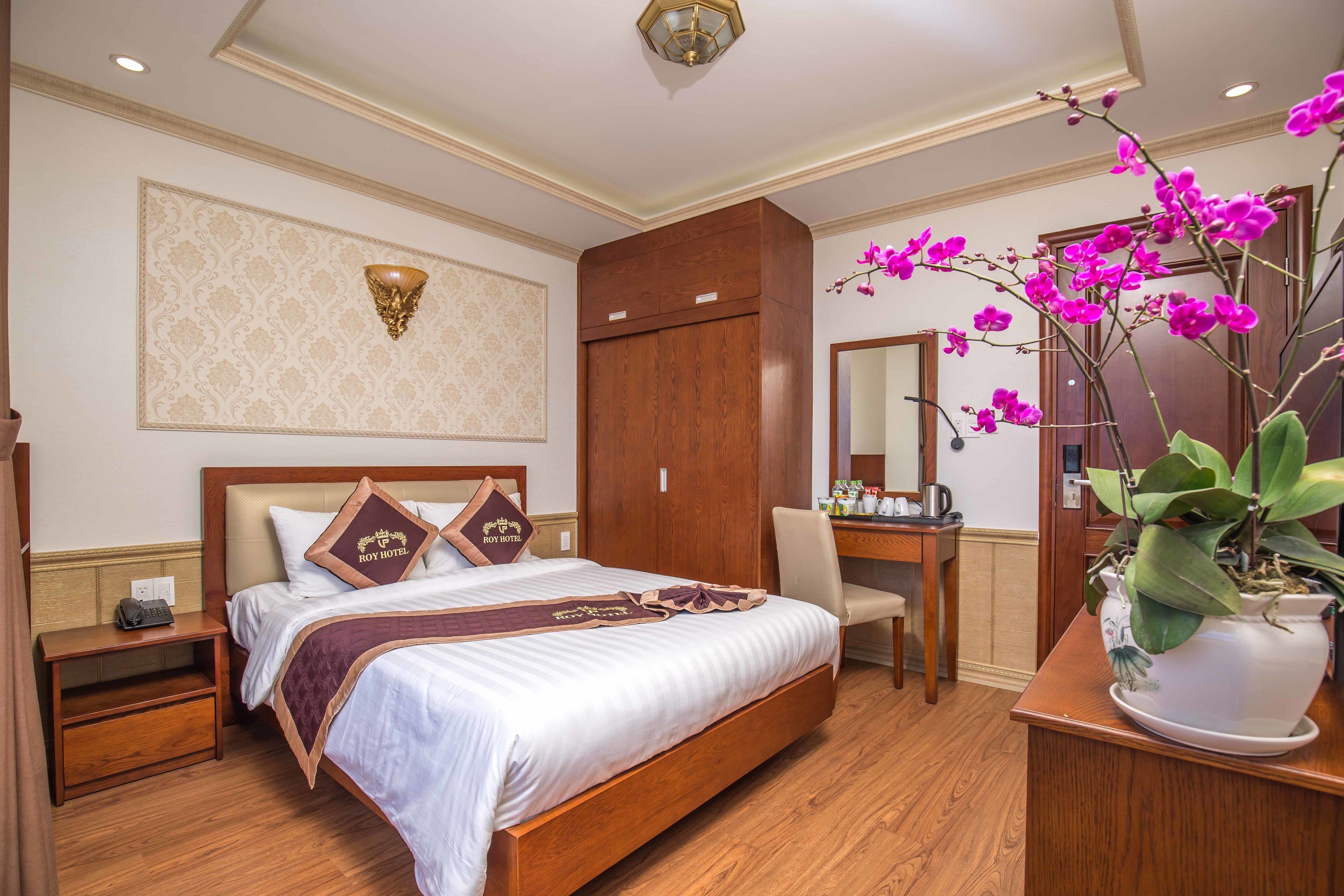 Une chambre d’hôtel avec un grand lit, une armoire en bois, un bureau avec une chaise, une table de chevet, un miroir et une orchidée en pot.