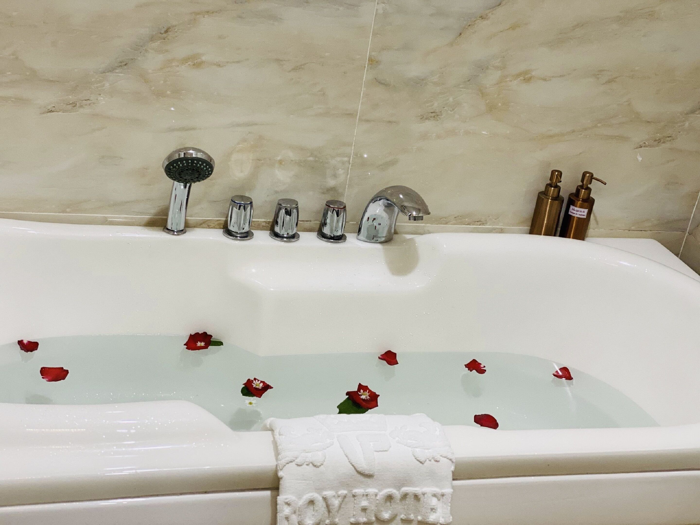 Une baignoire dans laquelle flottent des pétales de rose, une serviette ornée du mot « ROYAL » brodé dessus, et deux bouteilles de shampoing posées à côté.