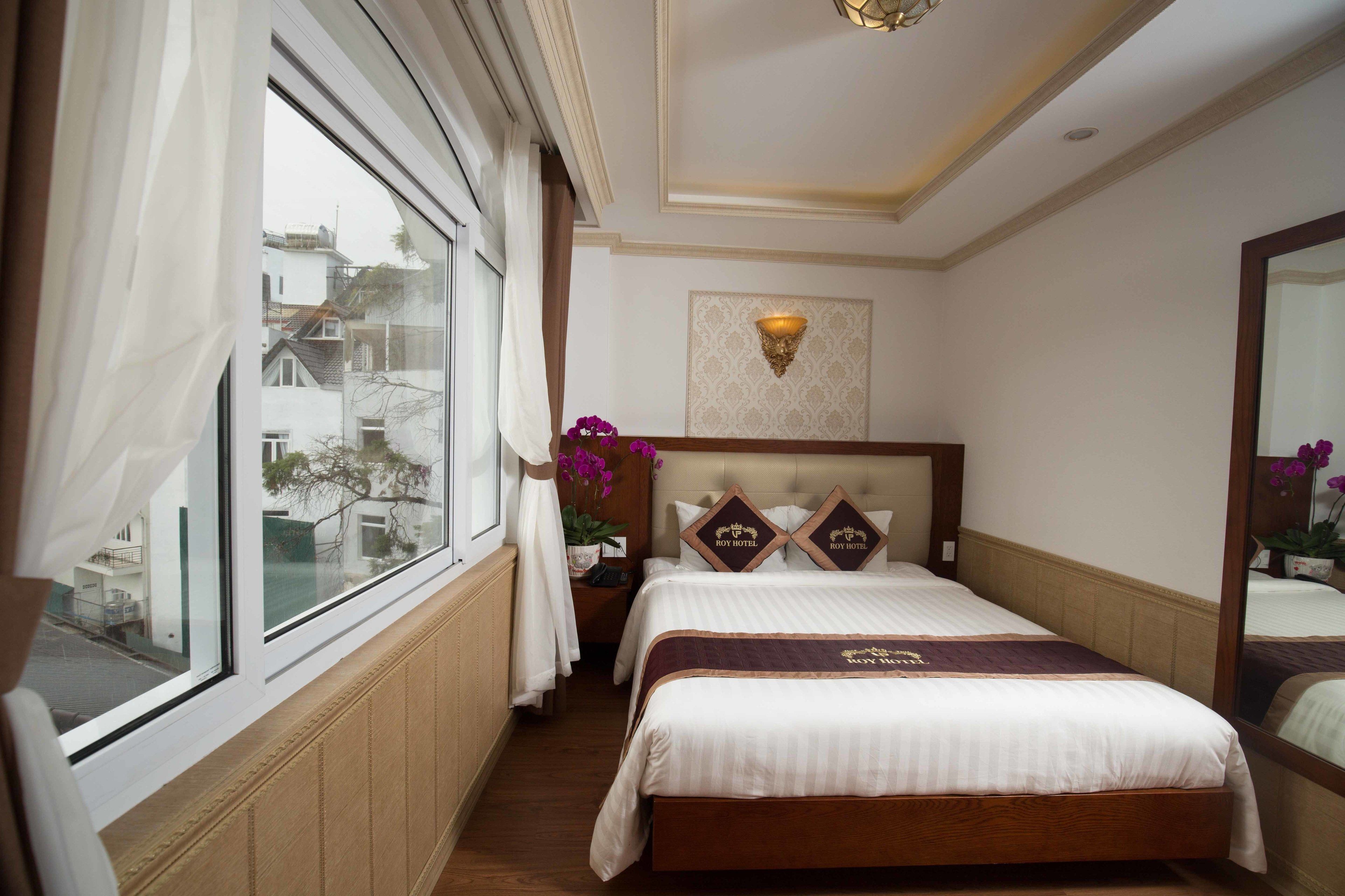 A hotel room with a large window, a bed with a headboard, a nightstand, and a mirror.
