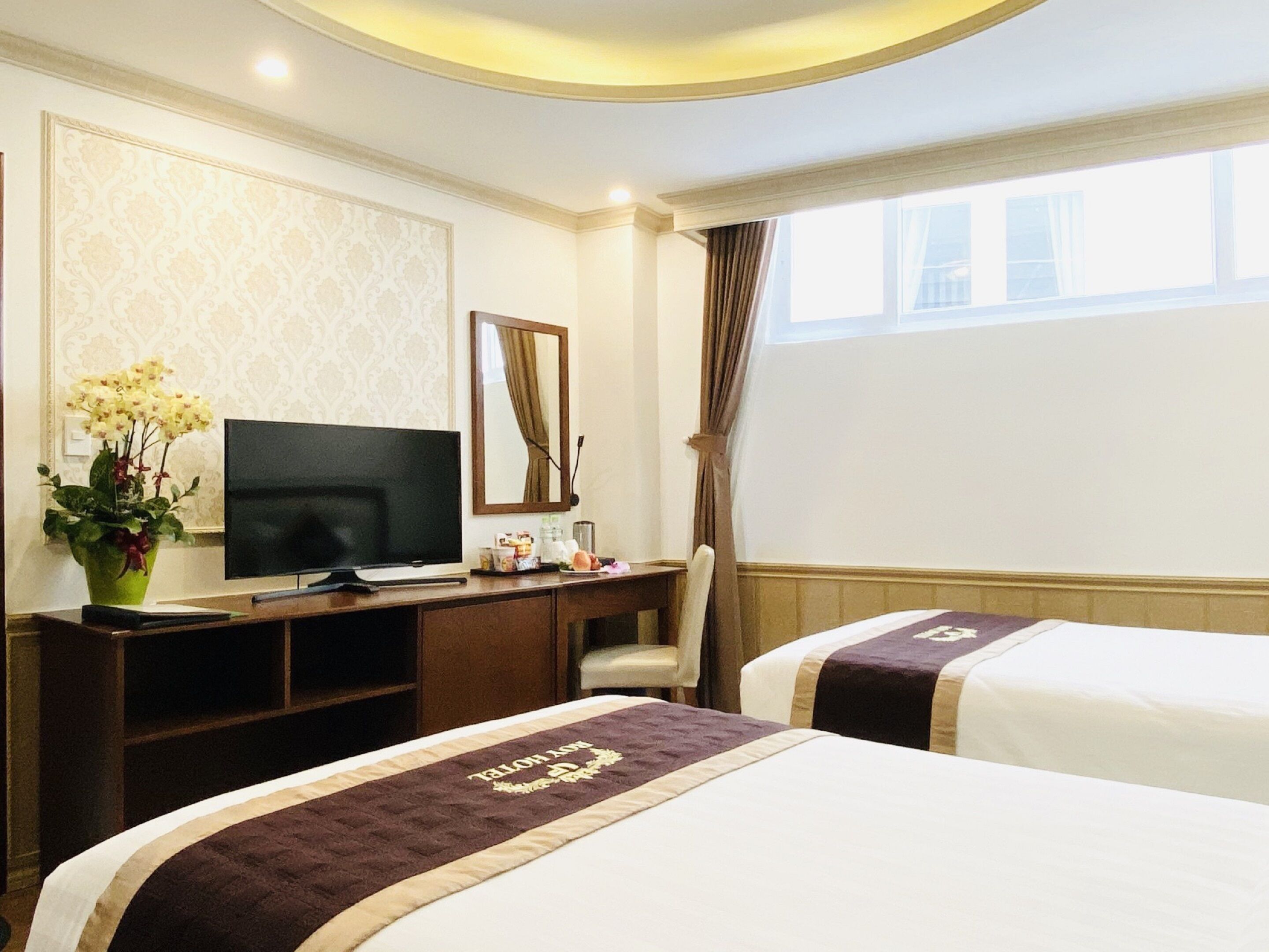 A hotel room with a bed, a desk with a TV, a chair, a window with curtains, and a wall with a patterned wallpaper.