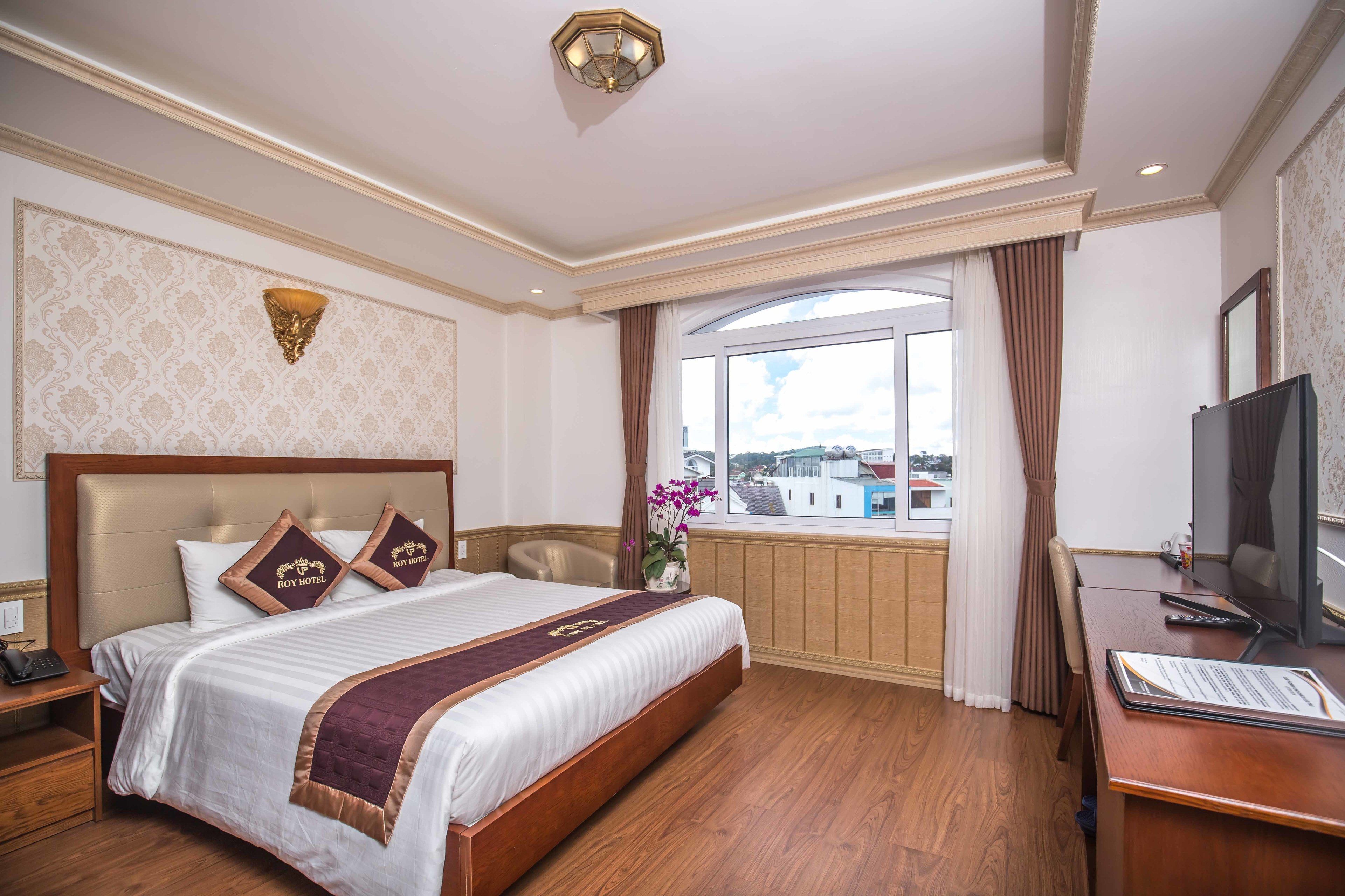 A hotel room with a large bed, a desk, a television, and a window with a view of buildings.