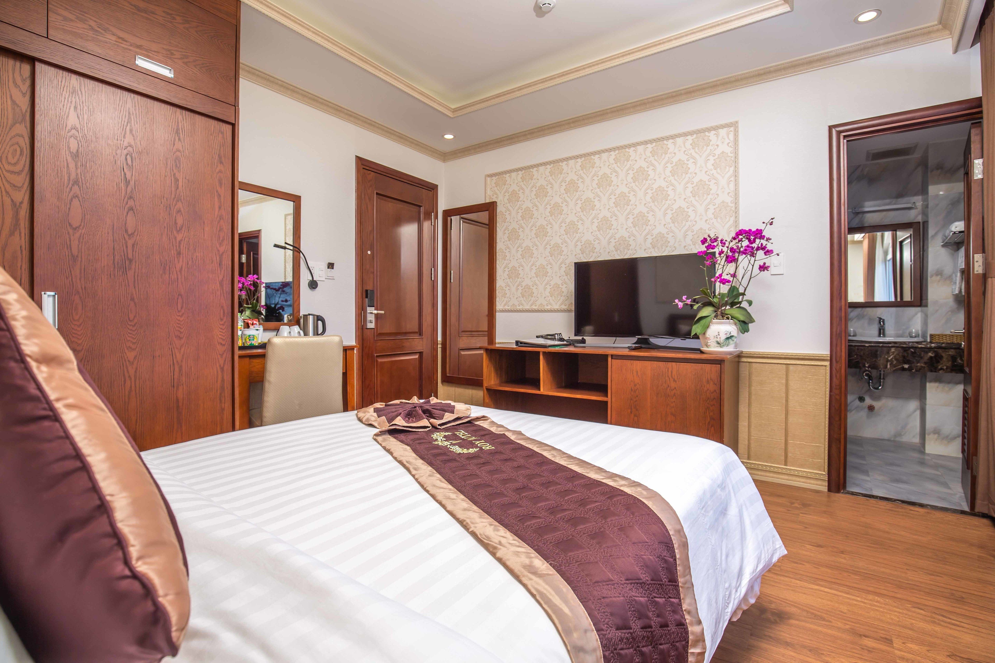 A hotel room with a large bed, wooden wardrobe, a desk with a computer, a TV, and a bathroom with a glass shower.