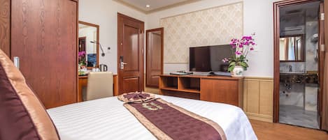 A hotel room with a large bed, wooden wardrobe, a desk with a computer, a TV, and a bathroom with a glass shower.